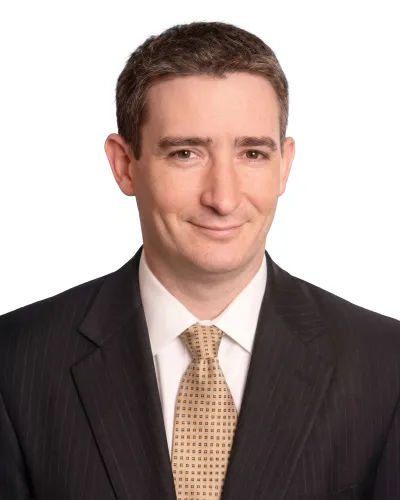 Professional headshot of a man in a dark suit and patterned beige tie smiling against a white background