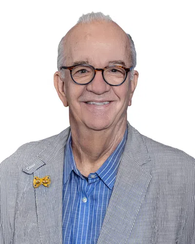 Smiling elderly man wearing round glasses, a blue striped shirt, and a gray pinstripe blazer with a yellow bow pin.
