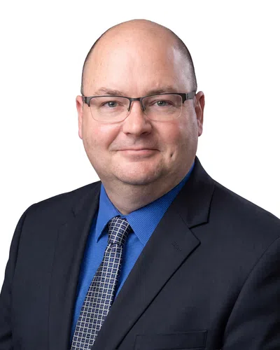 Middle-aged man wearing glasses, blue shirt, patterned tie, and black suit jacket smiling against white background