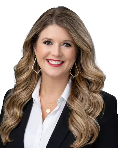 Smiling woman with long wavy blonde hair wearing gold earrings, white blouse, and black blazer on white background