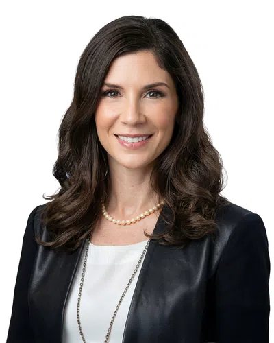 Smiling professional woman with wavy dark hair wearing a black leather blazer, white blouse, and pearl necklace.