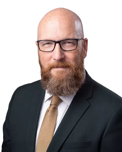 Bald man with glasses and a thick beard wearing a black suit and gold tie on white background