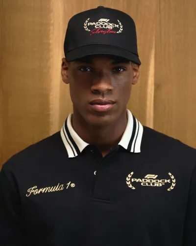 Man wearing black Formula 1 Paddock Club polo shirt and cap with embroidered logos against wooden background