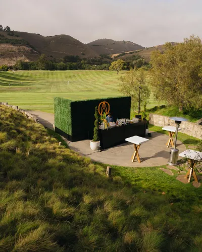 Outdoor bar setup with greenery backdrop and cocktail station overlooking a golf course and hills under cloudy sky