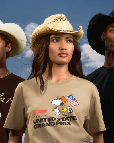 Three models wear Western hats and Peanuts-themed United States Grand Prix t-shirts under a blue sky.