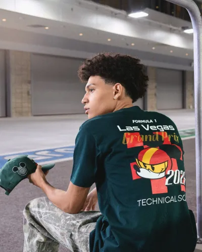 Young man in casual wear sitting near metal railings in an urban setting holding a green cap