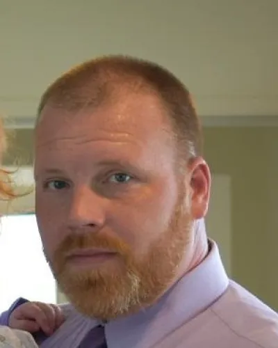 Close-up of a red-haired bearded man in a lavender shirt holding a child indoors with neutral background.