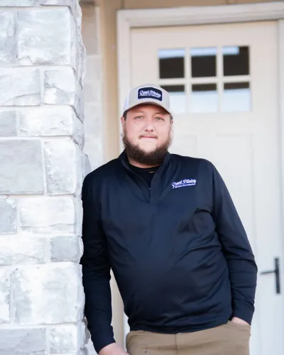 Man in a black long-sleeve shirt and cap stands relaxed by a brick column outside a house.