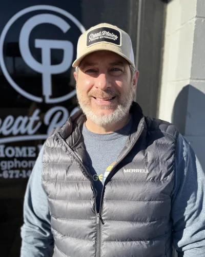 Smiling man wearing Great Plains Home Exteriors cap and Merrell vest standing outside company storefront
