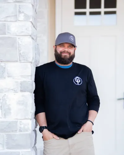 A man in a black shirt and cap stands by a stone wall, smiling confidently at the camera.