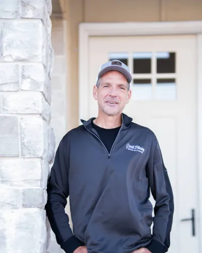 A man in a gray polo stands next to a stone column and door, smiling confidently at the camera.