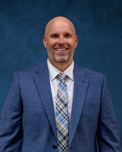 Smiling bald man in a blue plaid suit jacket with a white shirt and patterned tie against a blue textured background.