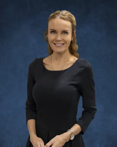 Smiling woman with blonde hair in a black dress standing against a solid blue textured background.
