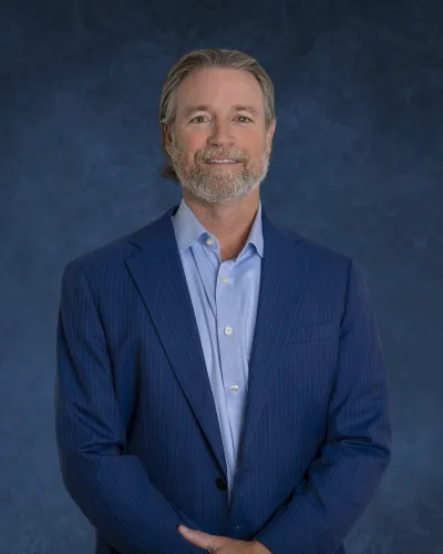 Middle-aged man with gray hair and beard wearing a blue suit and light blue shirt against a dark blue background