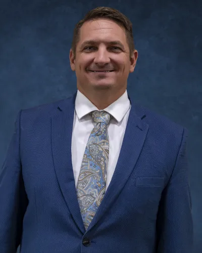 Smiling man in a blue suit, white shirt, and patterned tie against a dark blue background