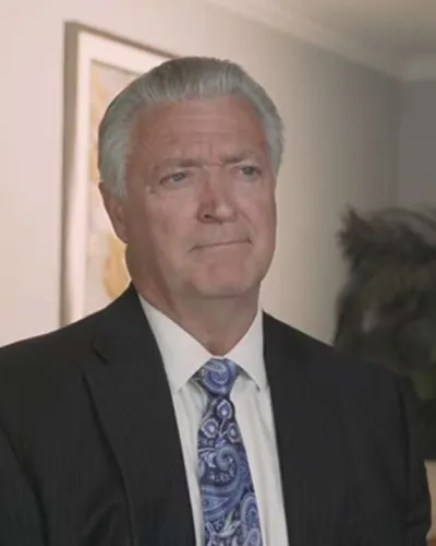 Man with light beard wearing a blue pinstripe suit and light blue shirt standing against a dark blue textured background