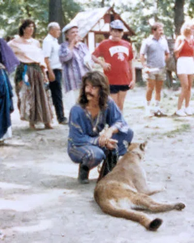 Group of people outdoors around a man kneeling beside a large mountain lion resting on the ground in a wooded area.
