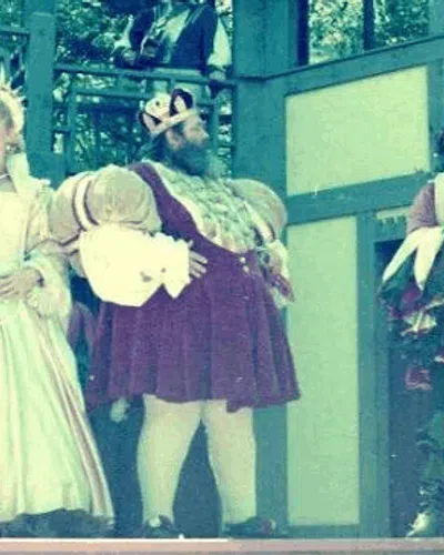 Actors in elaborate Renaissance costumes perform on an outdoor stage with a Tudor-style set design.