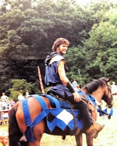 Smiling man in medieval costume on a horse with blue and white barding at an outdoor event.