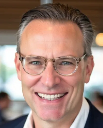 Smiling middle-aged man with glasses and gray hair in a blurred indoor setting, wearing a suit jacket.