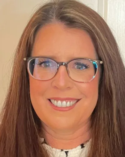 Smiling woman with long brown hair and glasses posing indoors against a light-colored wall.