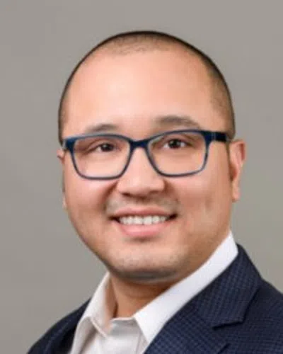 Smiling man wearing blue glasses and a checkered navy suit jacket over a white shirt against a gray background