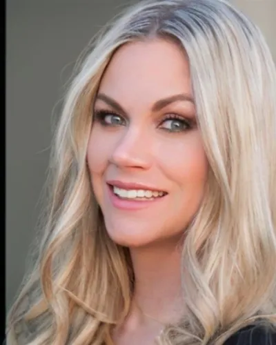 Close-up portrait of a smiling woman with long blonde hair and blue eyes in soft natural light.