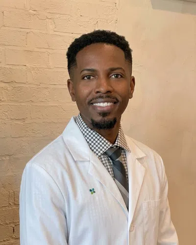 Smiling male doctor wearing white coat, checkered shirt, and striped tie against a light brick wall background.