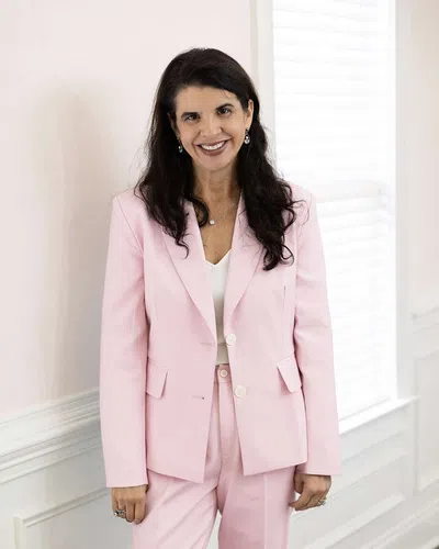 Smiling woman with long dark hair wearing a light pink suit and white blouse standing indoors by white window blinds.