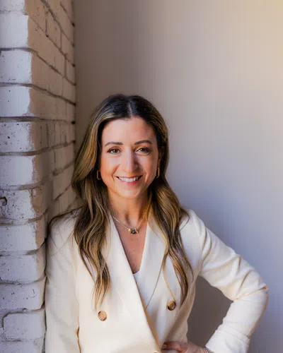 Smiling woman in white blazer leaning against white brick wall with hand on hip and long hair.
