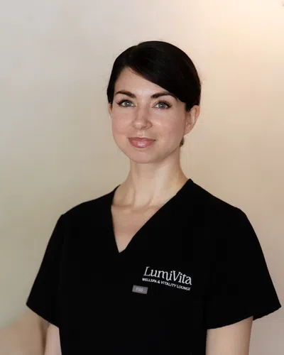 Woman in black LumiVita Wellspa & Vitality Lounge uniform with neutral background smiling gently