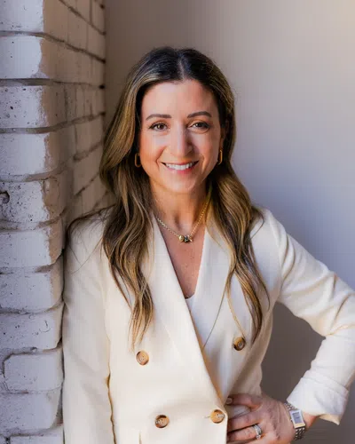 Smiling woman in white blazer leaning against white brick wall with hand on hip and long hair.