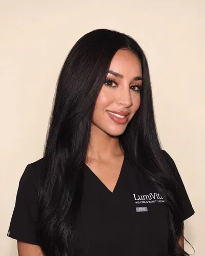 Smiling woman with long black hair wearing black LumiVita Wellspa uniform against a cream background.