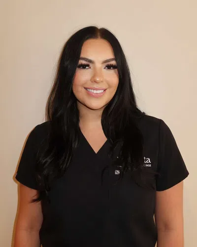 Smiling woman with long dark hair in black medical scrubs standing against a beige background.