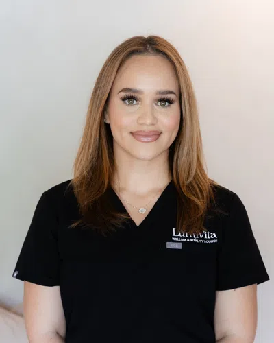 Smiling woman with light brown hair wearing a black LuLa Vita wellness lounge uniform standing against a light background.