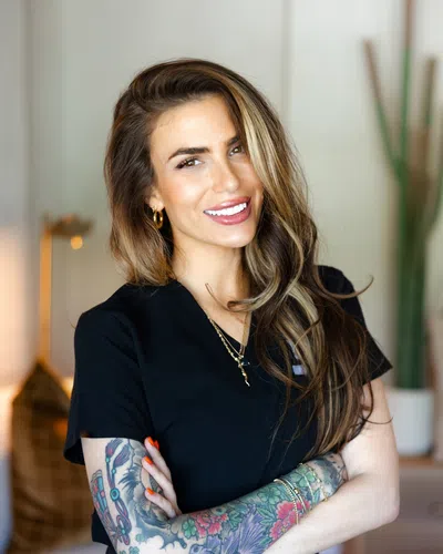 Smiling woman with long hair and colorful tattoos wearing black scrubs with arms crossed indoors.