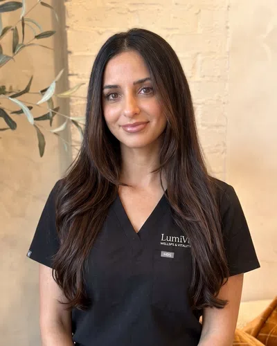 Smiling woman with long dark hair wearing black medical scrubs standing indoors with a plant and brick wall background.