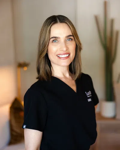 Smiling woman in black medical scrubs standing in a warmly lit room with blurred background elements.