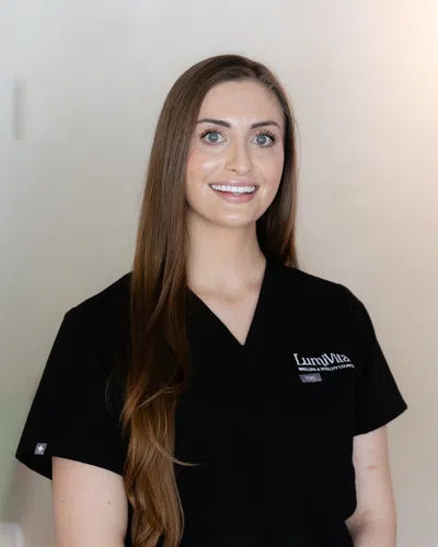 Smiling woman with long brown hair wearing black LumiVida scrubs against a neutral background.