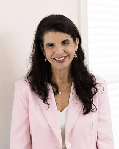 Smiling woman with long dark hair wearing a light pink suit and white blouse standing indoors by white window blinds.