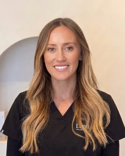 Smiling woman with long blonde hair wearing black scrubs stands before a neutral background with a small niche.