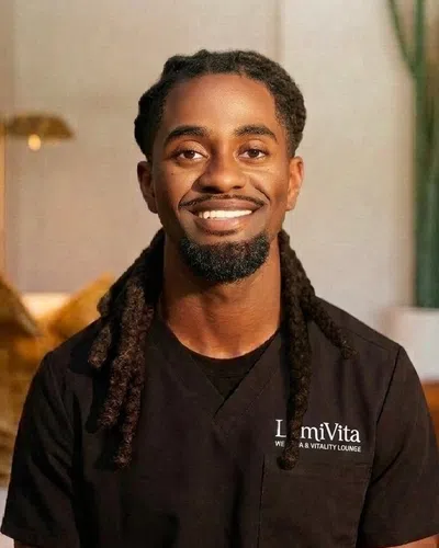 Smiling man with dreadlocks wearing a black shirt in a cozy room with plants and warm lighting.