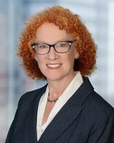 Professional woman with curly red hair, glasses, navy blazer, white blouse, and necklace smiling in office setting