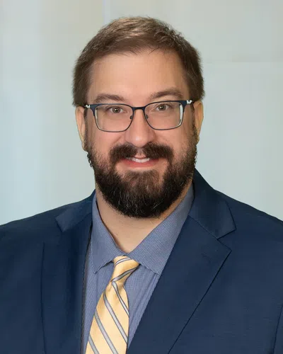 Smiling man with glasses, beard, blue suit, checkered shirt, and yellow striped tie against plain background