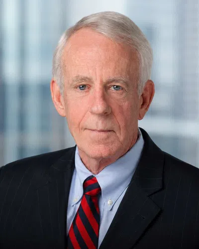Older man with white hair wearing a black suit, blue shirt, and red striped tie with a blurred office background