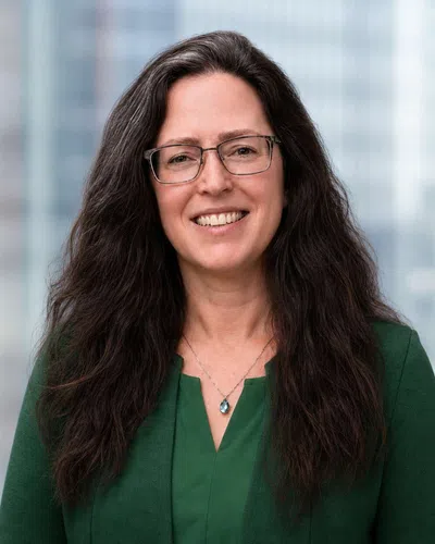 Smiling woman with long dark hair wearing glasses and green top with a teardrop pendant necklace against blurred cityscape.