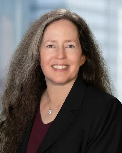 Smiling woman with long gray hair wearing black blazer and pendant necklace against blurred urban background