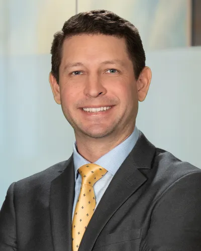 Smiling man in black suit, light blue shirt, and yellow patterned tie posing with crossed arms indoors.