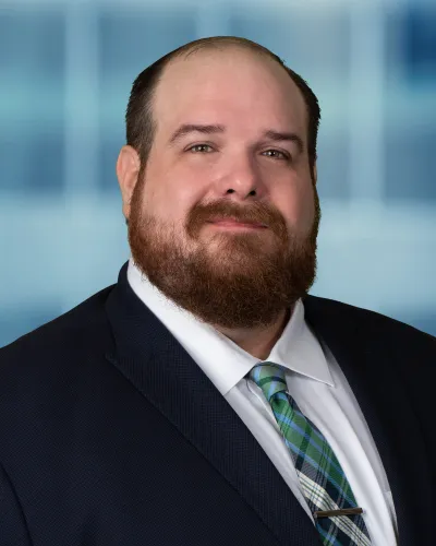 Smiling bearded man in a dark suit with a green plaid tie against a blurred blue background.