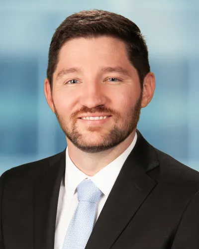 Smiling man with short brown hair and beard wearing a gray plaid blazer and light blue shirt on gray background